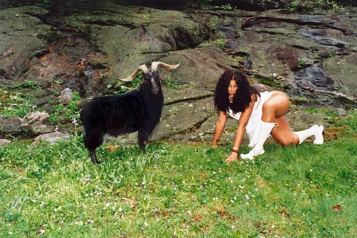 SZA crouching on grass wearing white dress and boots beside a large black goat against a rocky background for Interview Magazine