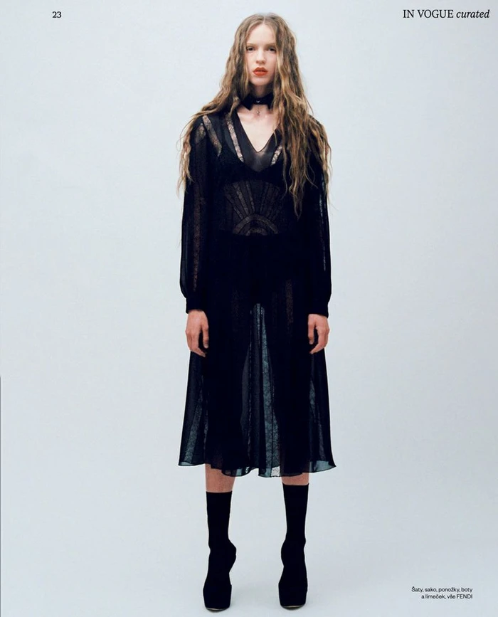 Model with long wavy hair wearing a sheer black dress and black boots against a white studio background