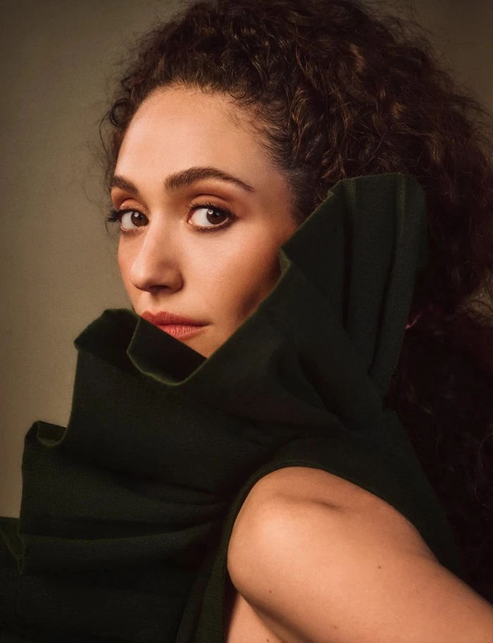 Close-up portrait of Emmy Rossum with curly dark hair wearing a high-collared dark green garment against a neutral background