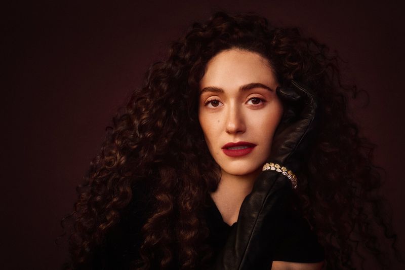 Emmy Rossum with voluminous dark curly hair, wearing a black leather glove and sparkling bracelet against a dark background