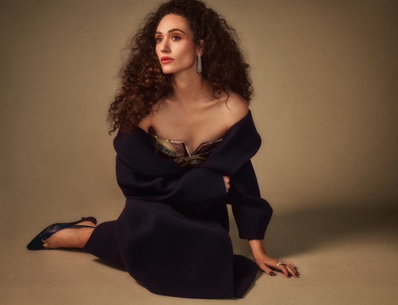 Emmy Rossum wearing a dark coat and strapless top seated against a warm textured studio backdrop