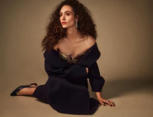 Emmy Rossum wearing a dark coat and strapless top seated against a warm textured studio backdrop