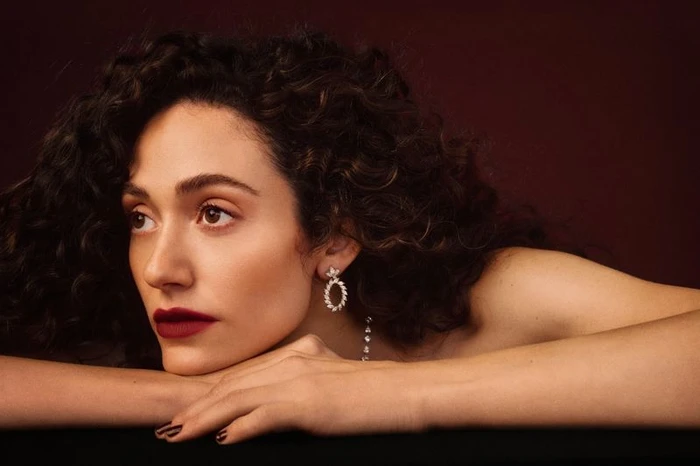 Emmy Rossum resting her chin on crossed arms, wearing dark crimson lipstick and jeweled drop earrings against a dark background