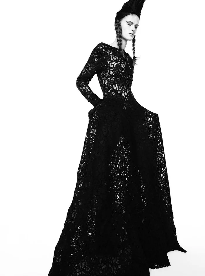 Lucia Richard in a long black lace dress with braids and a sculptural headpiece against a white background