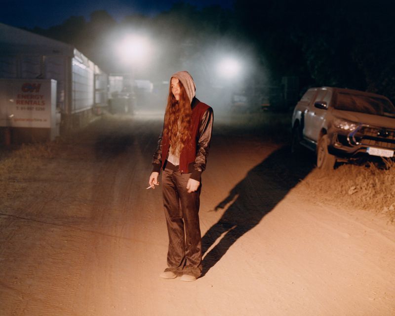 A person with long hair wearing a hooded varsity jacket standing on a dirt road at night under harsh lighting.
