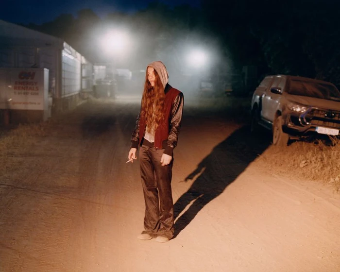 A person with long hair wearing a hooded varsity jacket standing on a dirt road at night under harsh lighting.