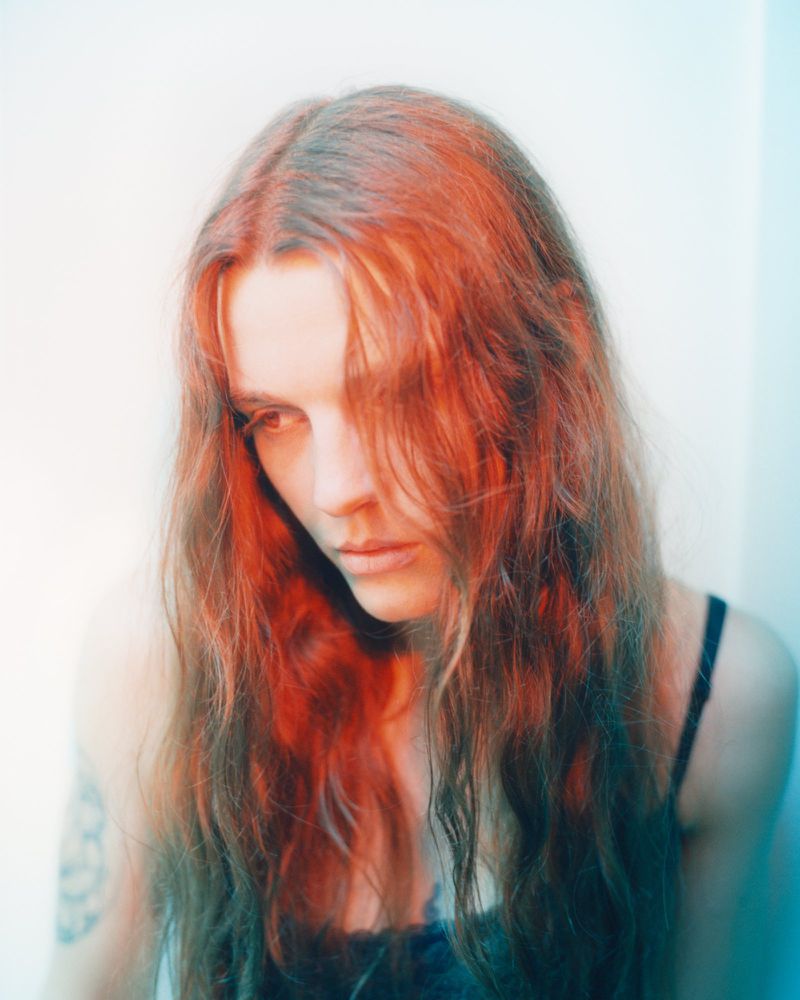Portrait of a woman with long red hair and dark top against a plain light background