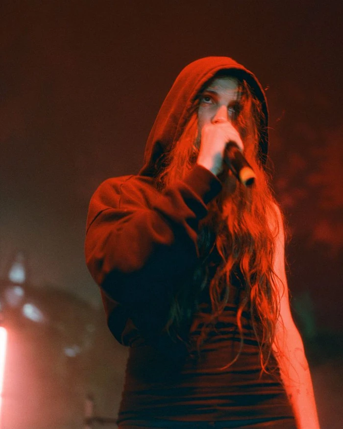 Performer in a red hoodie and dark top singing into a microphone under low red stage lighting.