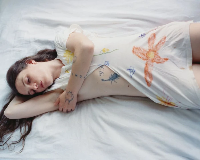 Woman with long dark hair lying on white bedsheets wearing a light-coloured dress with colourful crayon drawings