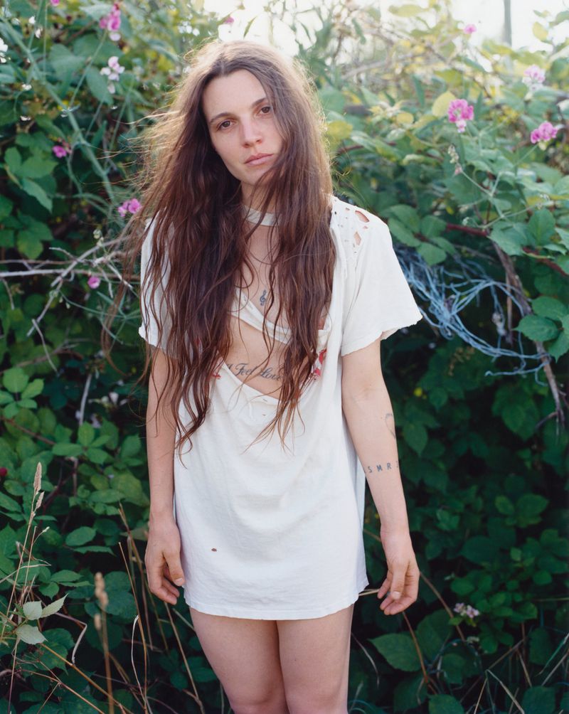 Woman with long brown hair wearing a white t-shirt dress standing outdoors amidst green foliage and pink flowers.