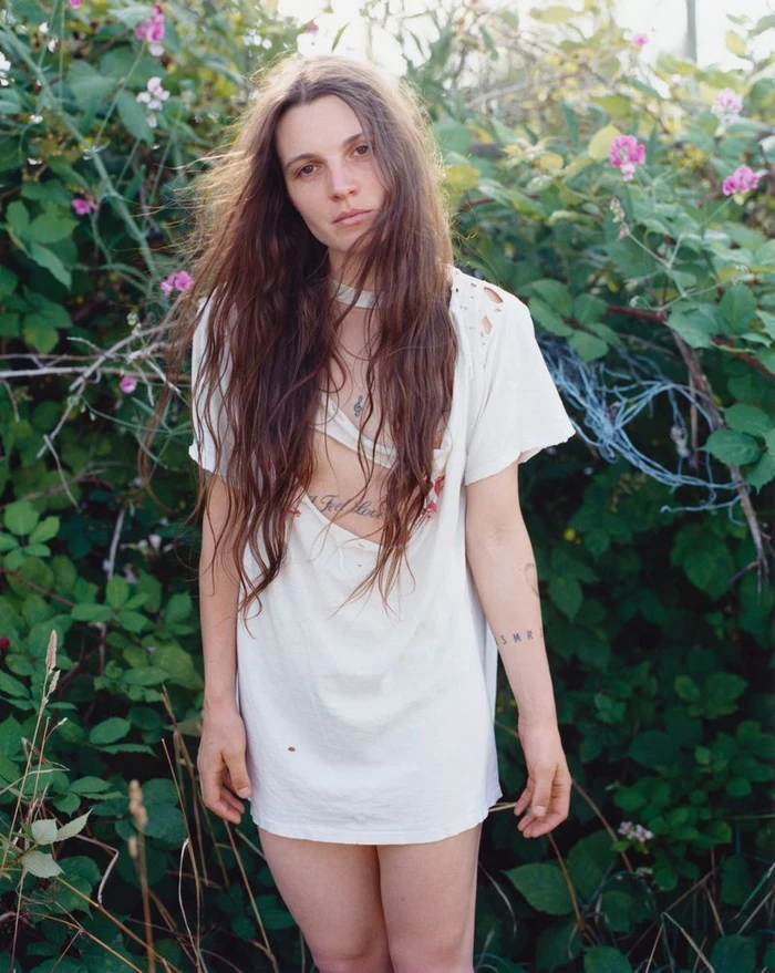 Woman with long brown hair wearing a white t-shirt dress standing outdoors amidst green foliage and pink flowers.