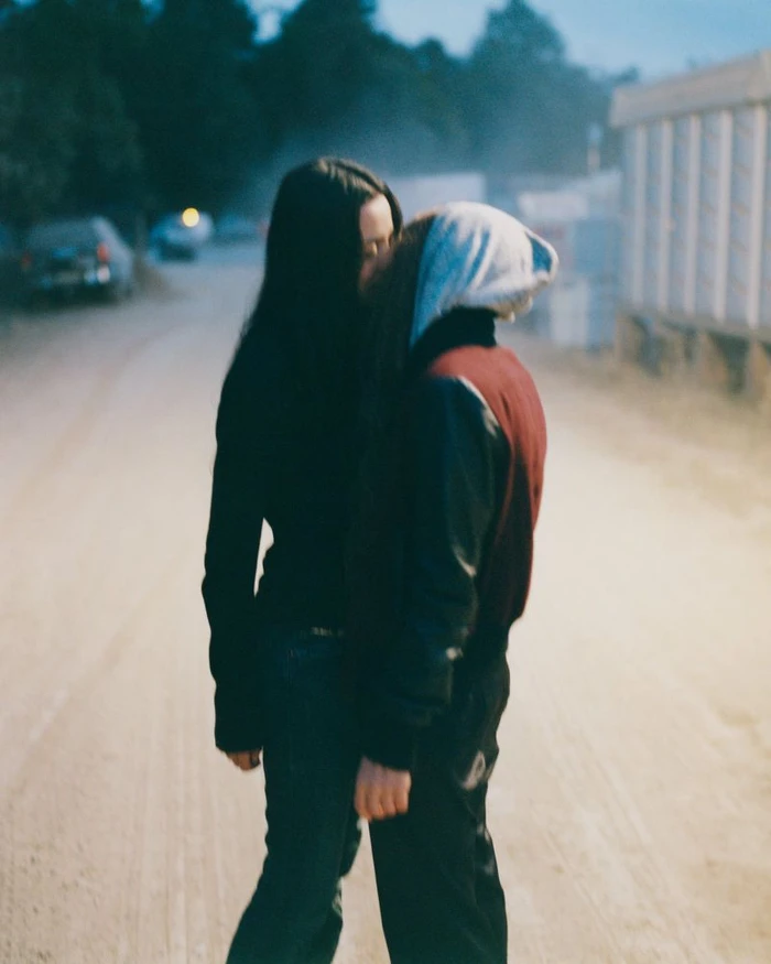 Two people standing together outdoors on a dirt road at dusk, with one wearing a dark jacket and jeans and the other in a hoodie.