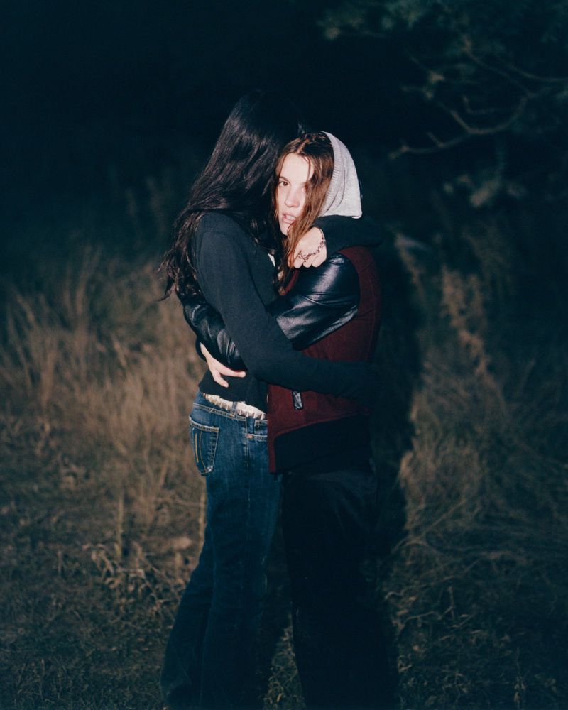 Two women embracing outdoors at night in a field with dark foliage background