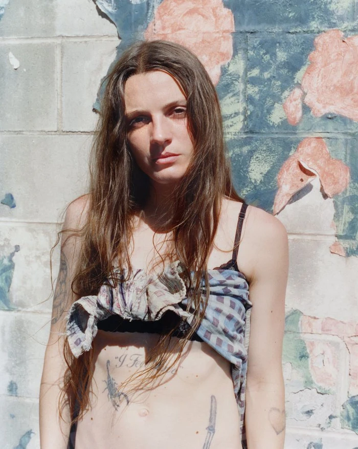 Portrait of a woman with long brown hair posing against a weathered wall with faded floral murals.