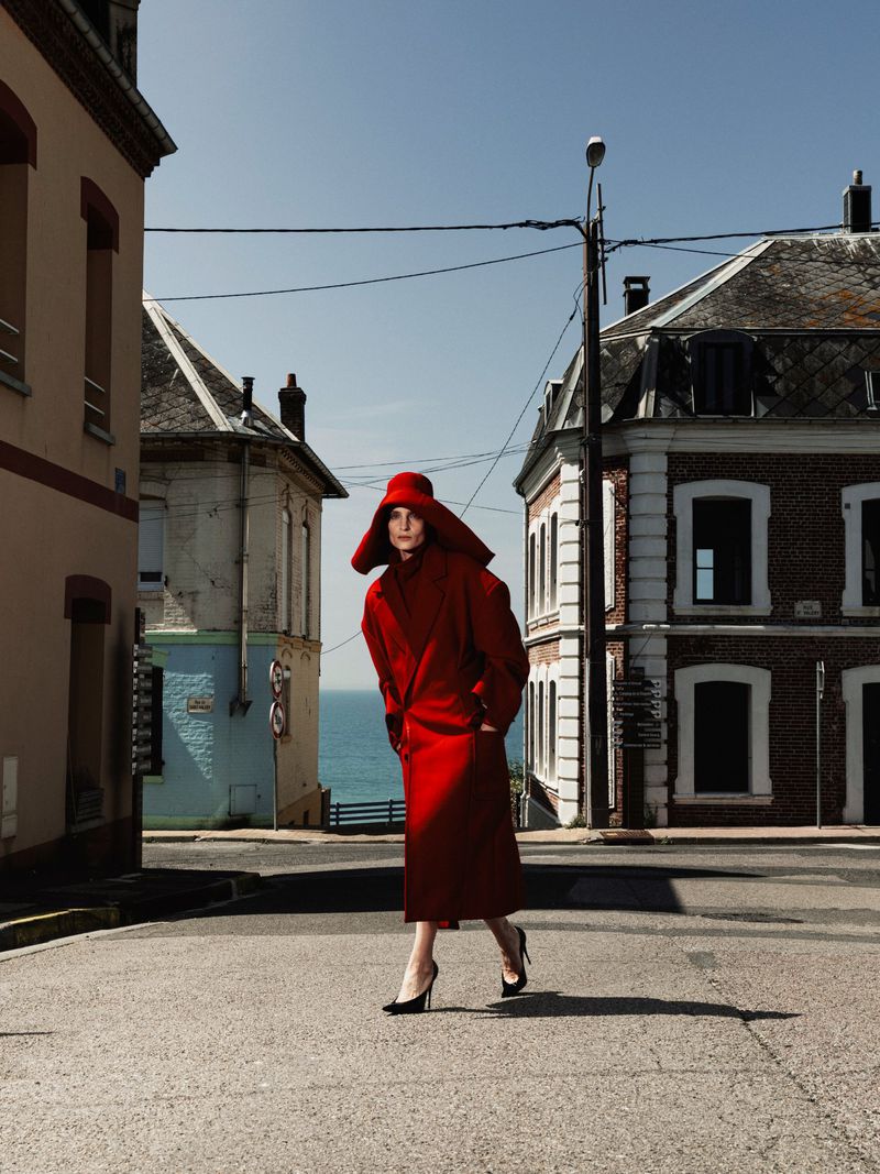 Model in a bright red coat and matching wide-brimmed hat walking down a sunlit coastal street between old European buildings