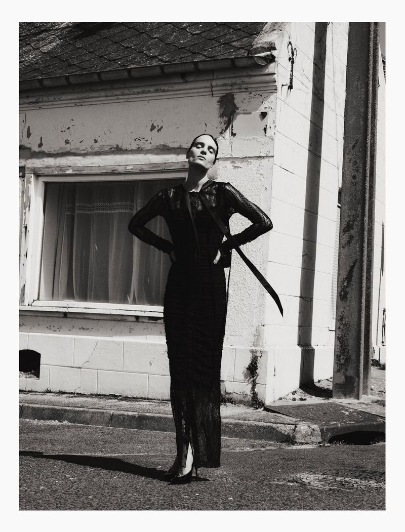 Model in a black lace dress standing with hands on hips against a rustic building exterior.