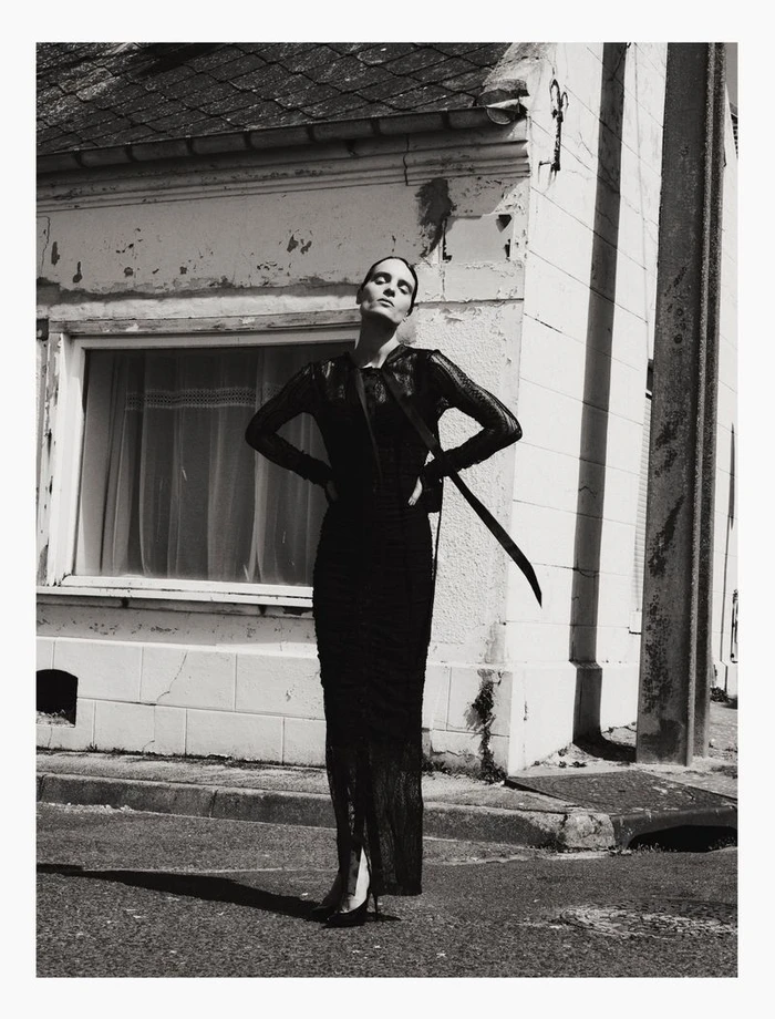 Model in a black lace dress standing with hands on hips against a rustic building exterior.