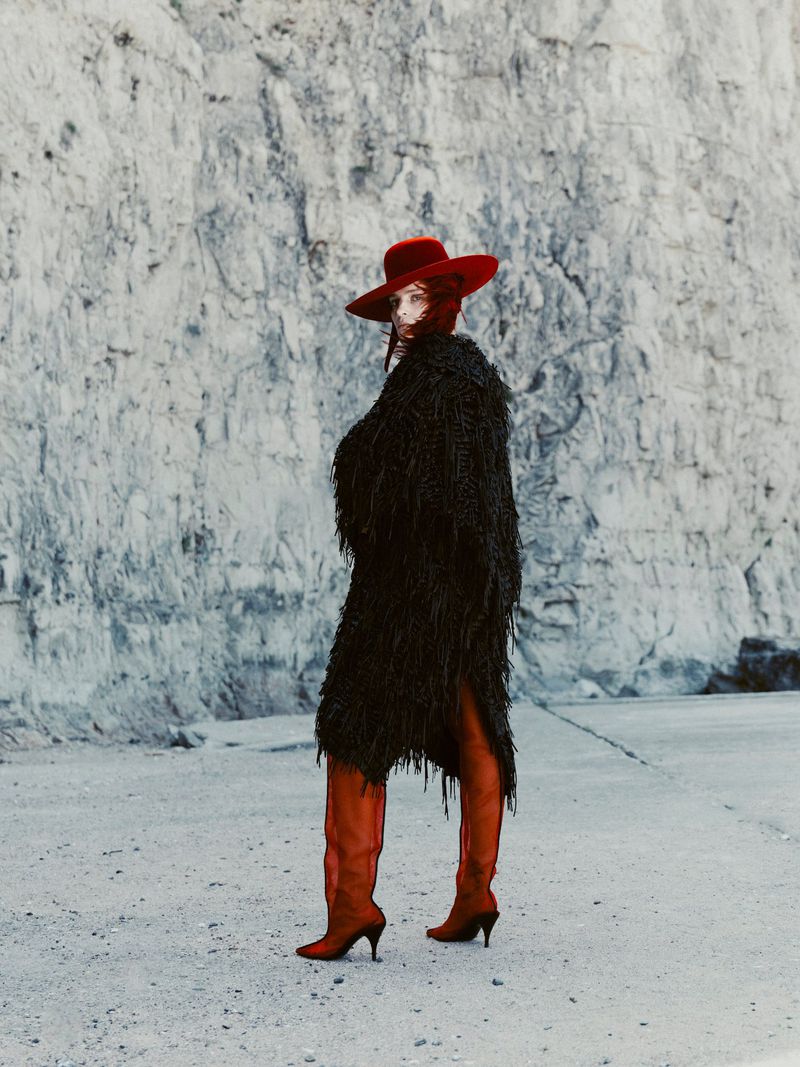 Model in a black textured coat and red hat standing before a rocky cliff face for Mode Metieres editorial by Tarek Mawad