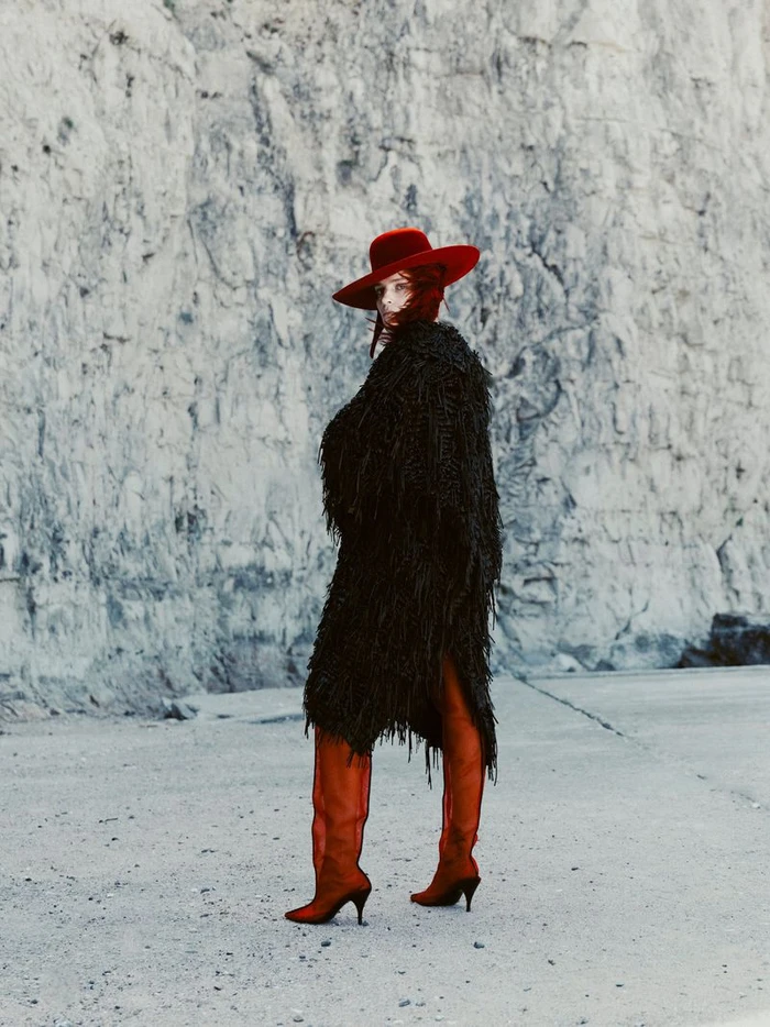 Model in a black textured coat and red hat standing before a rocky cliff face for Mode Metieres editorial by Tarek Mawad