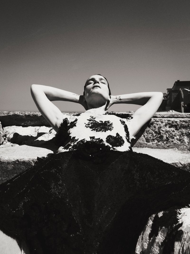 Model lying back with hands behind head on stone steps outdoors in strong sunlight, wearing a patterned top and dark skirt.