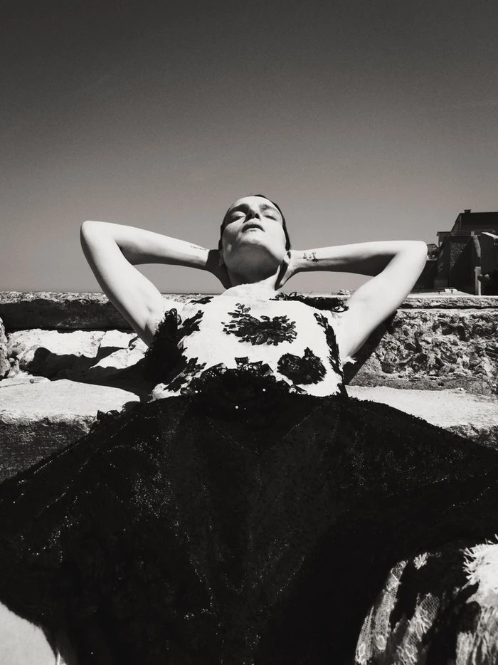 Model lying back with hands behind head on stone steps outdoors in strong sunlight, wearing a patterned top and dark skirt.