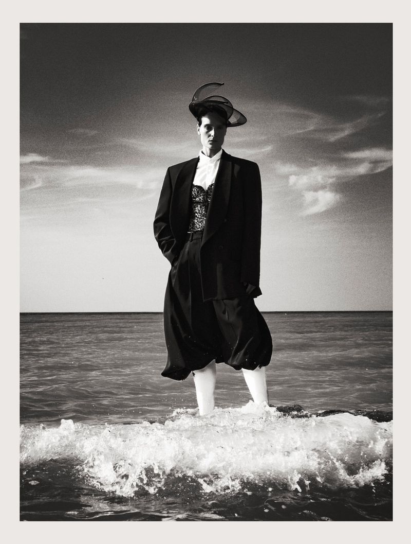 Model standing in ocean surf wearing a black blazer, white shirt, and sculptural hat under a cloudy sky.