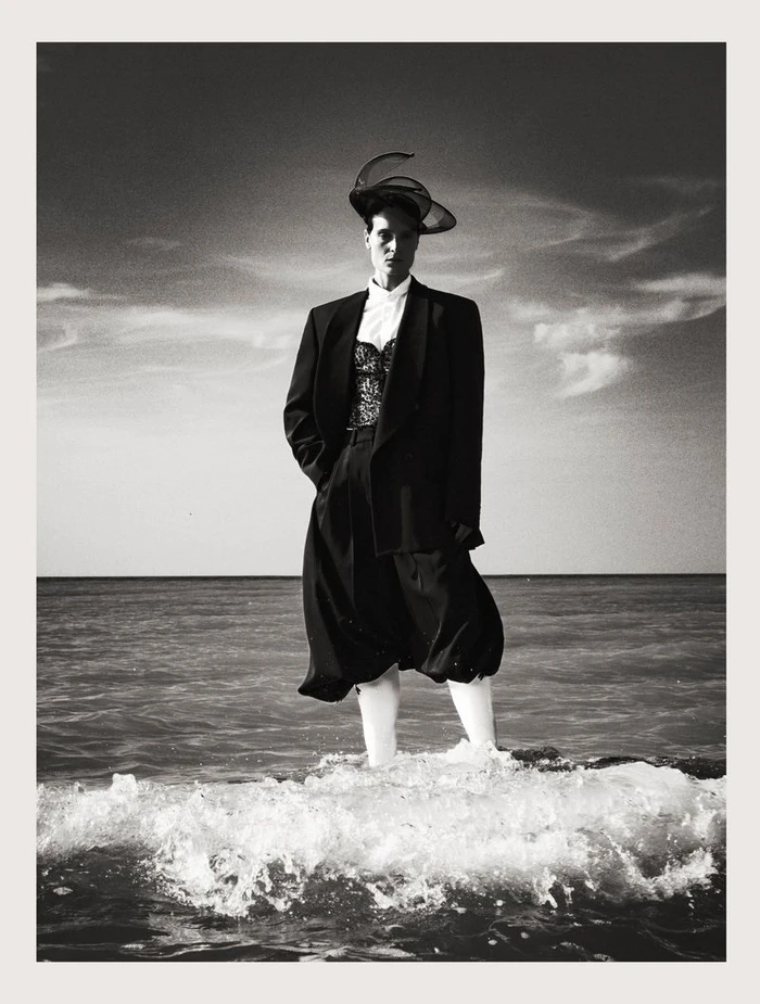 Model standing in ocean surf wearing a black blazer, white shirt, and sculptural hat under a cloudy sky.