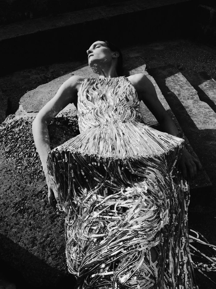 Model lying on stone steps wearing a heavily textured silver fringe dress under harsh sunlight