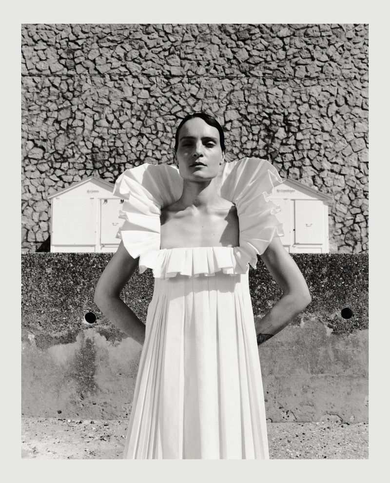 Model in a white pleated dress with a dramatic ruffled collar standing before a stone wall