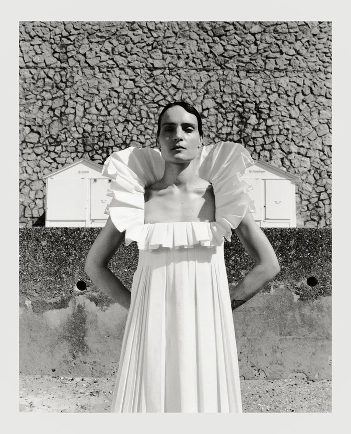 Model in a white pleated dress with a dramatic ruffled collar standing before a stone wall
