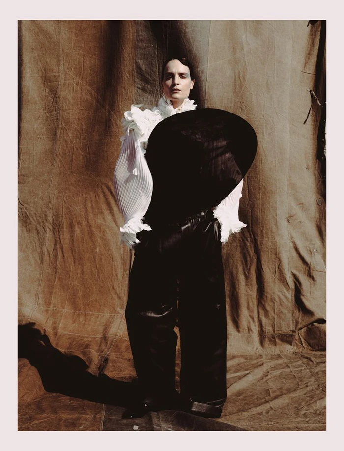 Model wearing a pleated white ruffled blouse and black trousers holding a large round black hat against a textured brown fabric backdrop