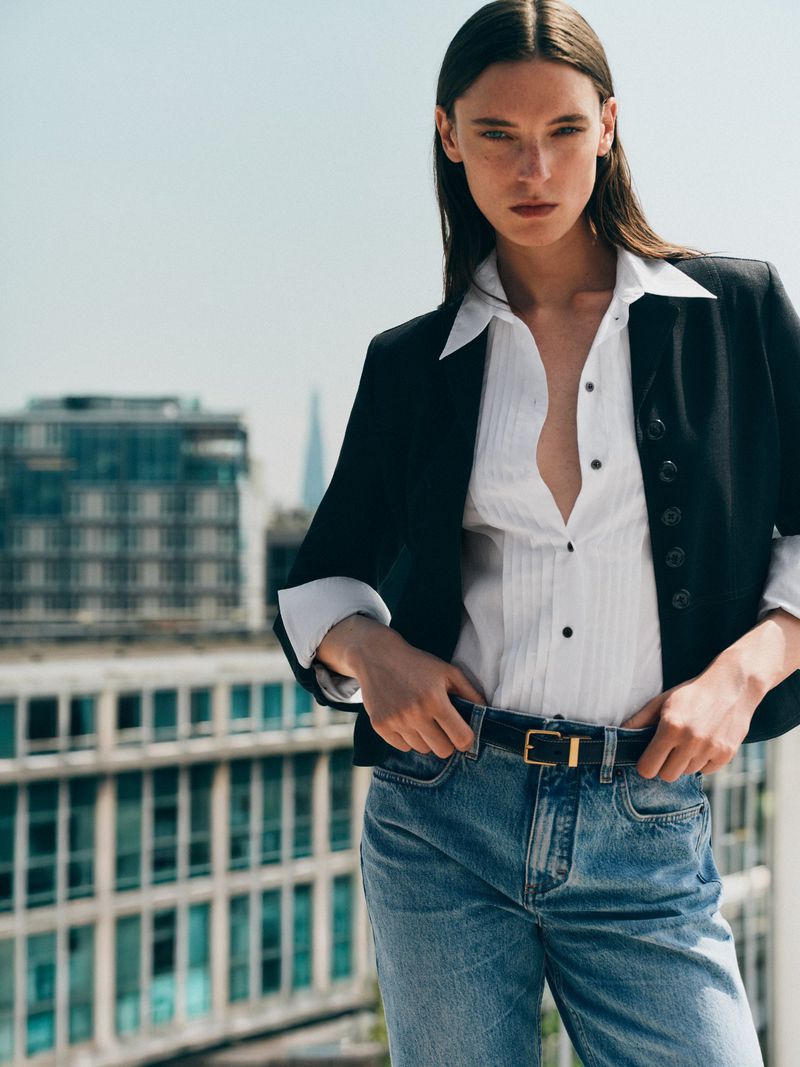 A fashionable woman with long brown hair poses in jeans, a white top, and a black jacket against a city backdrop, exuding a chic, modern style.