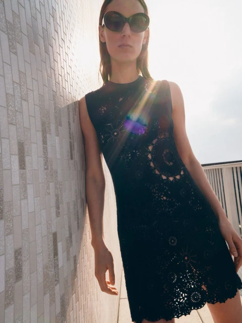 A woman stands beside a wall in a black lace dress and sunglasses, posing in the sunlight, showing off fashion and style.