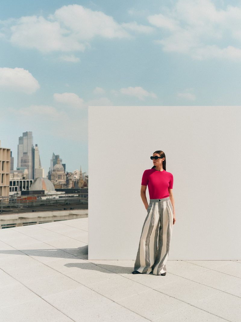 Woman in stylish outfit poses on rooftop with city skyline.  Fashion editorial shot.  Bright colors, sunny day. Wide-leg pants are dominant.