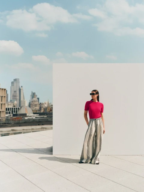 Woman in stylish outfit poses on rooftop with city skyline.  Fashion editorial shot.  Bright colors, sunny day. Wide-leg pants are dominant.