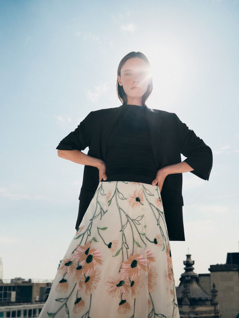 A woman poses in a black top and floral skirt against a rooftop and blue sky. The sun shines from behind the model.