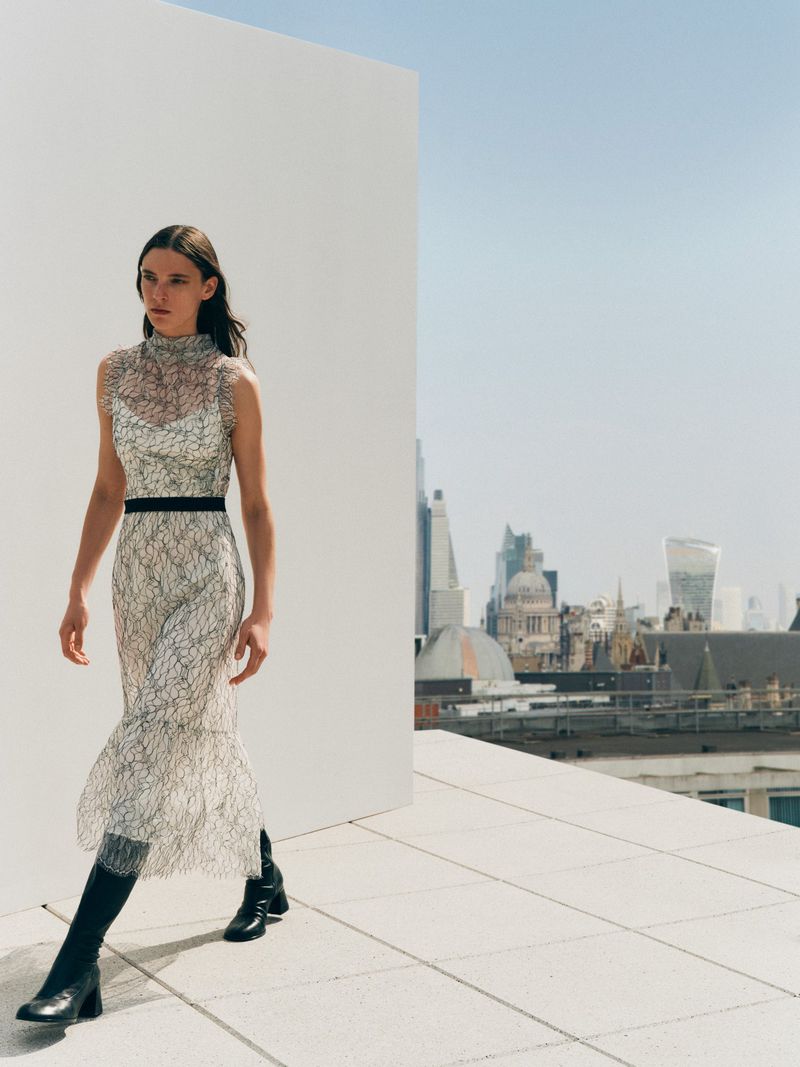 Fashion shot of a woman in an elaborate dress and black boots, walking on a rooftop with an urban cityscape in the background.