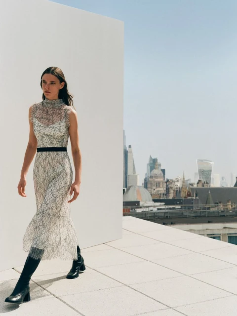 Fashion shot of a woman in an elaborate dress and black boots, walking on a rooftop with an urban cityscape in the background.
