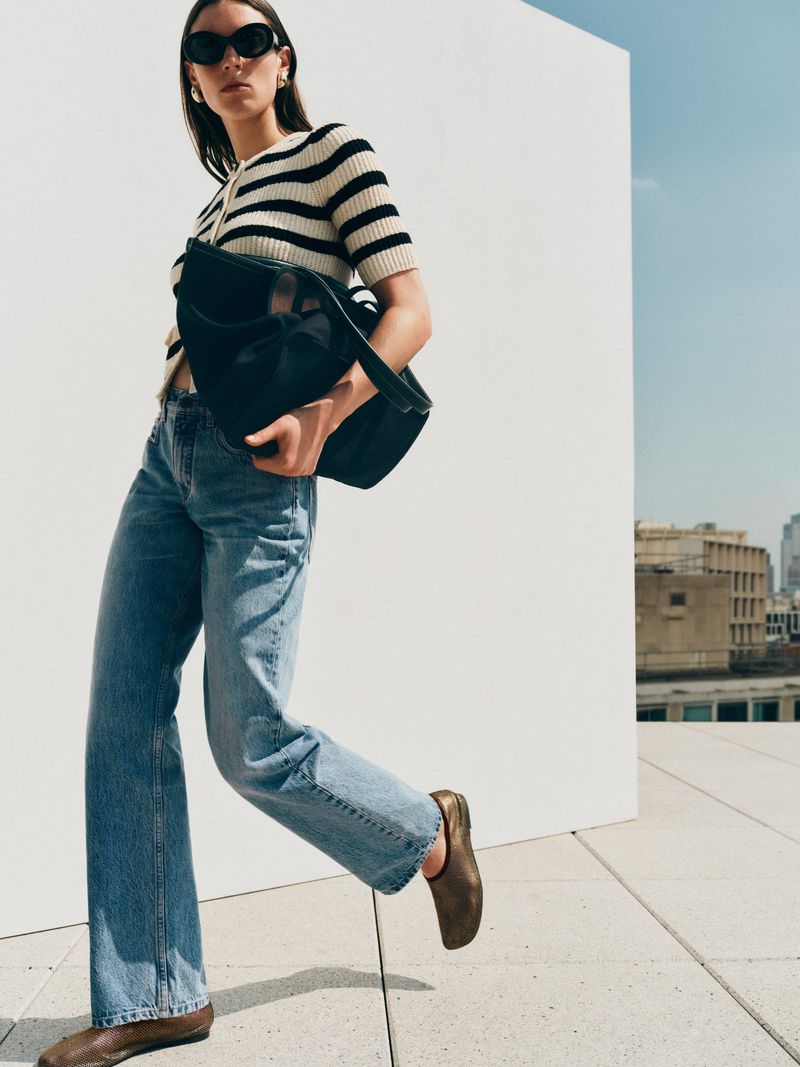 Stylish woman in jeans, striped top, and flats carrying a black bag walks on a rooftop with city buildings in the background.