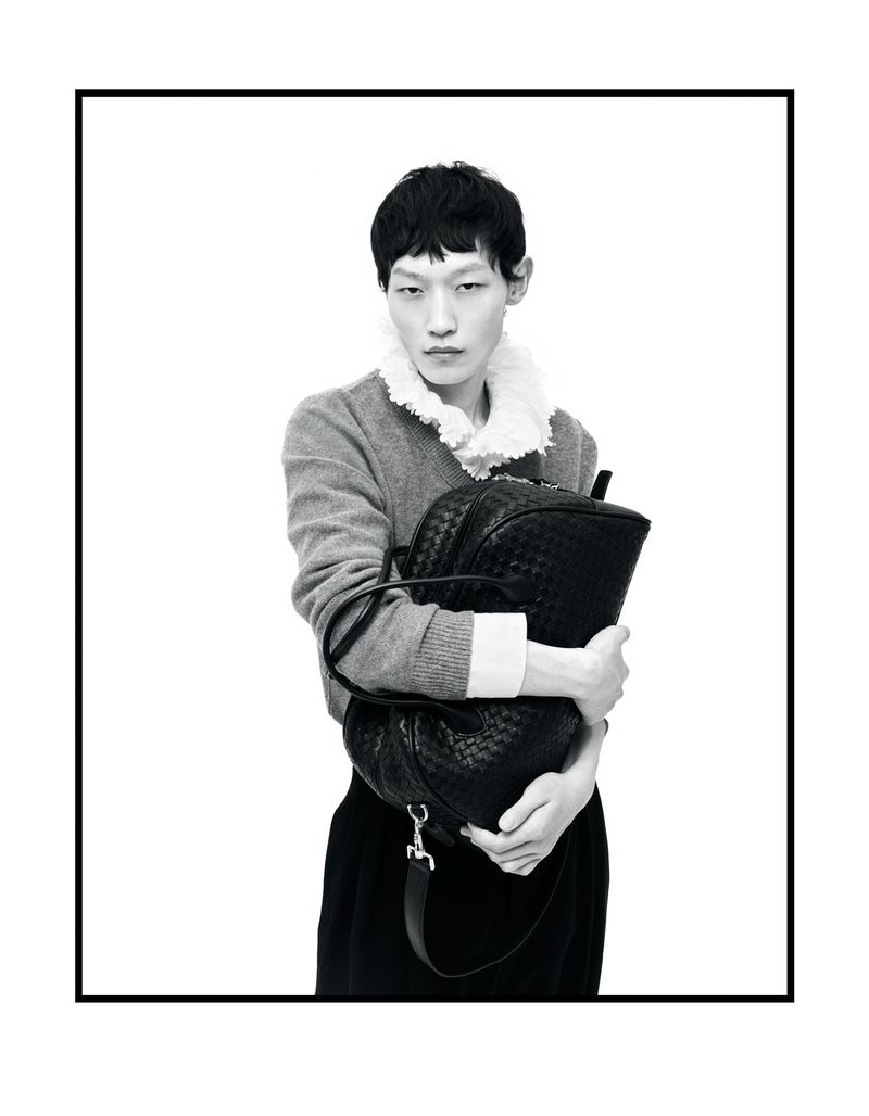 Man wearing a grey sweater with ruffled collar holding a black leather bag against a plain white background