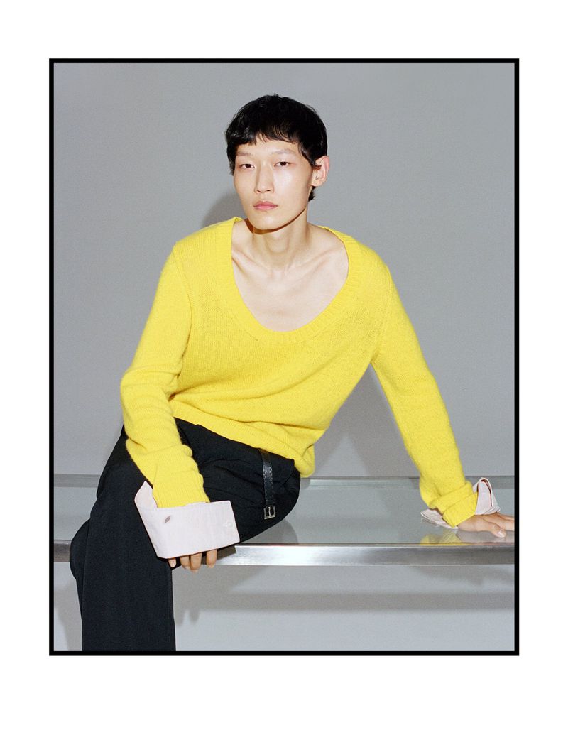 Asian male model wearing yellow sweater and black pants seated on a glass bench against a grey studio wall for Esquire China