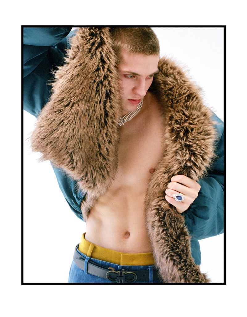 Close-up portrait of a male model wearing a furry collar jacket and pearl necklace against a plain white background