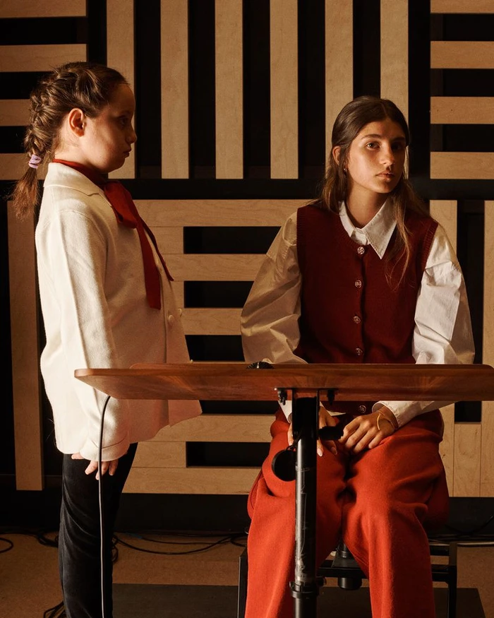 Two young girls posed in a wood-panelled interior, one standing in a white jacket and red scarf, the other seated in a red vest and trousers.