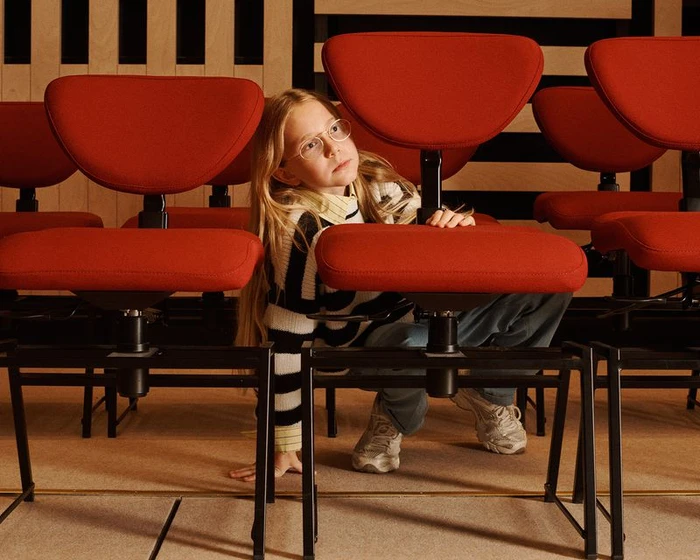 Child with glasses wearing striped sweater crouching among rows of red chairs