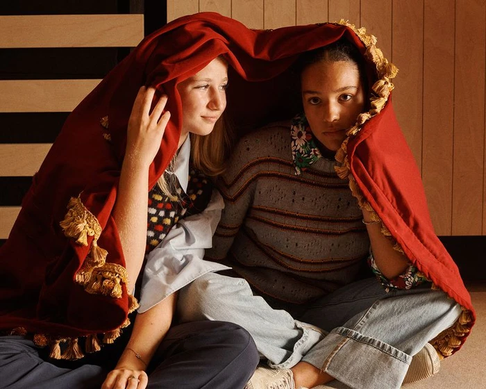 Two young models sitting together under a draped red fabric blanket with gold tassels for Vogue Italia.