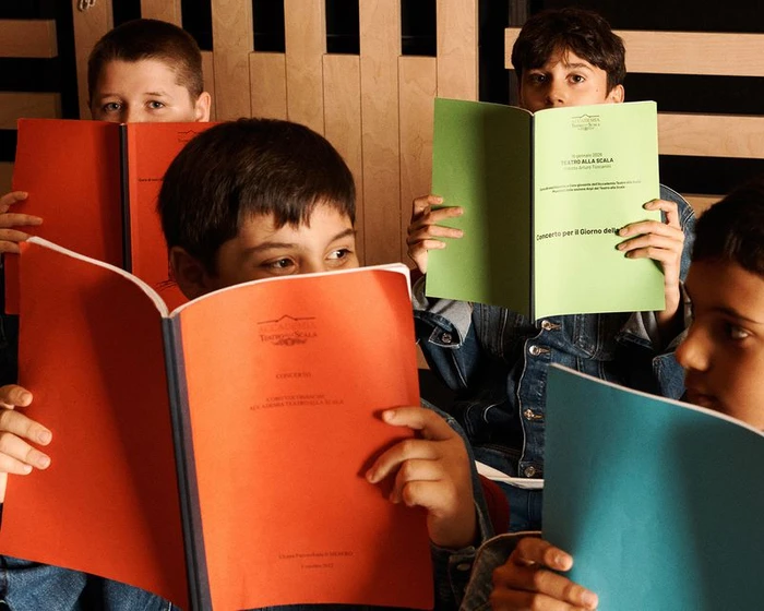 Four children holding up colourful booklets covering their faces in an indoor setting with a wooden slatted wall background.