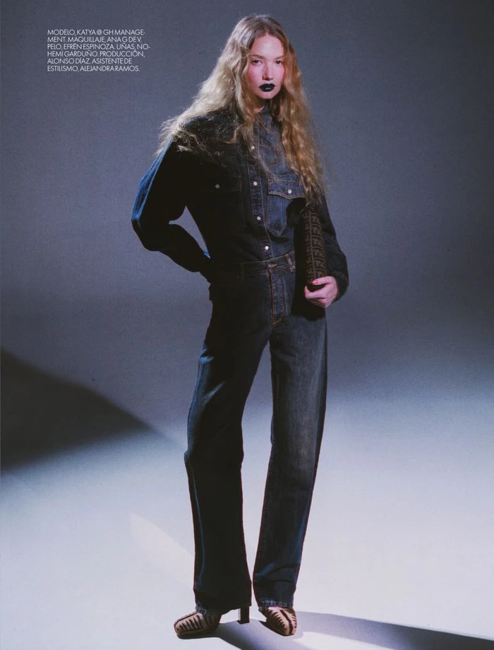 Model with long blonde wavy hair wearing a denim jacket and jeans against a dark studio background