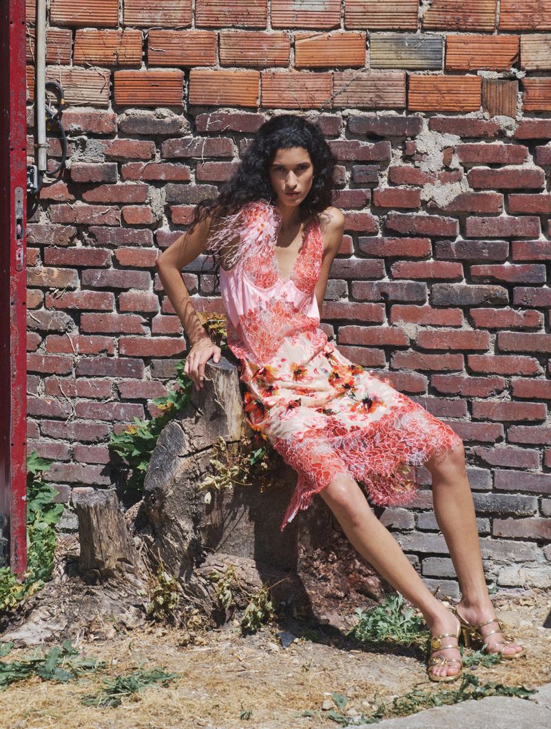 Model seated on a tree stump wearing a pink and orange patterned dress against a weathered brick wall.