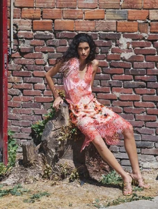 Model seated on a tree stump wearing a pink and orange patterned dress against a weathered brick wall.
