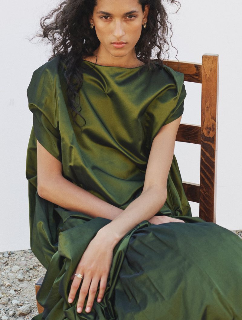 Model seated on a wooden chair wearing a lustrous green satin dress against a plain white background